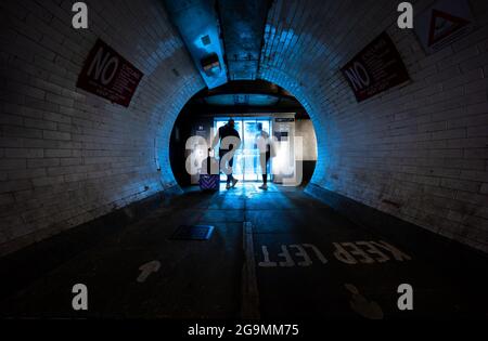 Pedestrian tunnel with tiled walls and ‘Keep Left’ floor markings as people walk toward a bright blue exit. Stock Photo