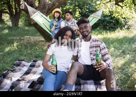 Positive black couple drinking beer and hugging while sitting on plaid outdoors. Two friends on background chatting in hammock. Young multiracial people having picnic. Stock Photo