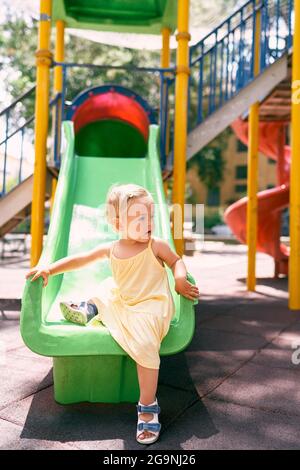 Little girl in a dress climbs down the childrens slide, turning her head to the side Stock Photo