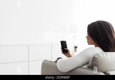 Home temperature, safety and control on mobile app. African american woman using smartphone, sitting on sofa, copy space Stock Photo