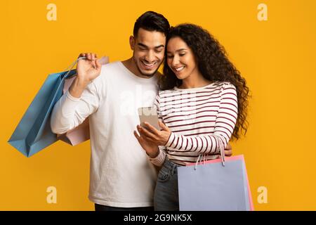 Discount App. Cheerful Arab Couple Holding Smartphone And Colorful Paper Shopping Bags Stock Photo