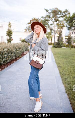 Smiling beautiful blonde teenage girl 17-18 year old wear stylish hat, leather bag and knit gray jumper walking on city street in park outdoors. Autum Stock Photo