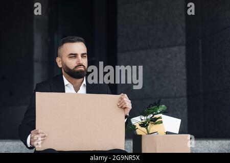 Upset fired male employee sitting outside and holding empty poster with box of personal belongings nearby Stock Photo