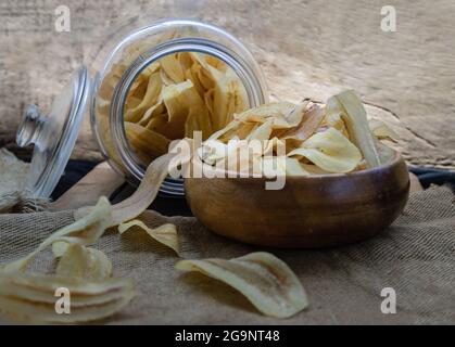 Homemade banana chips in wooden bowl and glass jar on sackcloth and old wooden background. Banana dessert, Long banana chips, Selective focus. Stock Photo