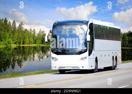 White coach bus travels on highway through rural scenery of a calm lake, green forest and blue sky with fairweather clouds on a day of summer. Stock Photo