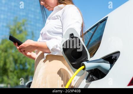 Woman plugging in the charger into her white electric car. Using a smartphone while waiting Stock Photo