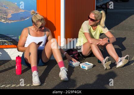 Two woman sitting on ground with fish & chips cartons in the evening at Poole Quay at Poole Harbour, Poole, Dorset UK in July Stock Photo