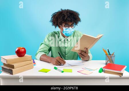 Black student guy wearing protective face mask and taking notes from book, sitting at table over blue background Stock Photo
