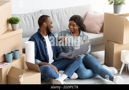 Cheerful Black Married Couple Sitting With Laptop Among Unpacked Cardboard Boxes Stock Photo