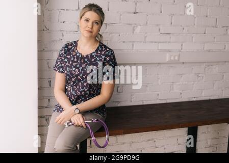 Medium shot portrait of attractive young woman in casual clothing sitting at desk holding stethoscope Stock Photo