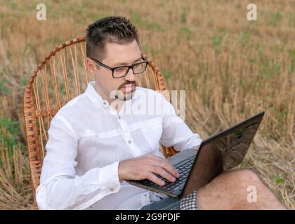 Young man with laptop sitting in a rocking chair outdoors. Handsome caucasian man with glasses working remotely outdoors with laptop. Creative remote Stock Photo
