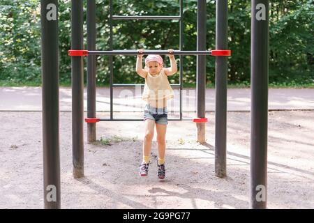 little girl pulls herself up on a horizontal bar in a city park on a summer day Stock Photo