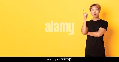 Portrait of thoughtful handsome asian man raising finger, suggesting idea, standing yellow background Stock Photo
