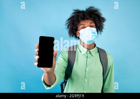 Black student guy in protective mask holding smartphone with blank screen in hands, standing on blue background Stock Photo
