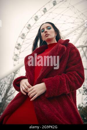 Pretty girl against the Ferris wheel. Retro (vintage) portrait of beautiful stylish young woman in amusement park, wearing red dress and fur coat Stock Photo