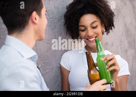 Multiracial couple of young lovers talking together holding and drinking bottles of beer. Shy African American woman with eyes closed. Stock Photo