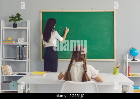 Schoolgirl sitting at desk and looking at teacher writing elementary sum on board Stock Photo