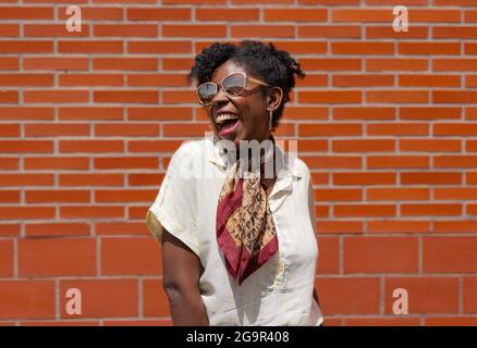 Delighted black woman in stylish clothes and sunglasses smiling and looking away while standing against brick wall on street