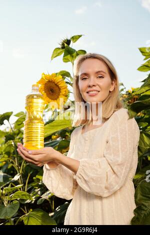 Gorgeous blond young woman holding bottle of sunflower oil while standing against large flowers in front of camera in the field Stock Photo