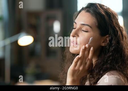 Young brunette pleased woman in silk pajamas applying natural cosmetic product on her face and enjoying it in the evening before sleep Stock Photo
