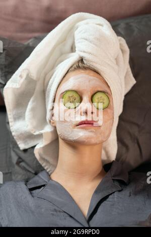Two girls in silk pajamas, towels on heads, clay masks on faces and cucumber slices on shut eyes lying on bed and enjoying beauty procedure Stock Photo