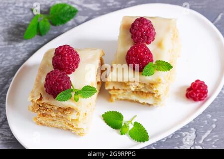 Pieces of Napoleon cake decorated with raspberries and lemon balm leaves on a light background. Stock Photo