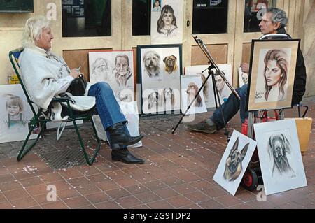 Candid shot of male street artist drawing the portrait of a mature Caucasian woman while smiling, smoking, talking and being surrounded by paintings Stock Photo