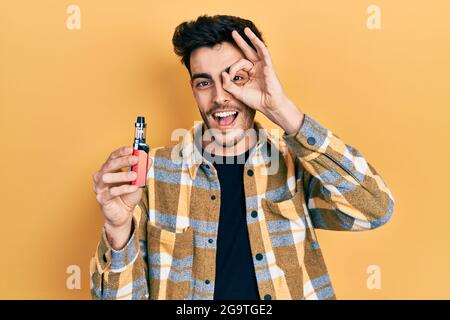 Young hispanic man holding electronic cigarette smiling happy doing ok sign with hand on eye looking through fingers Stock Photo