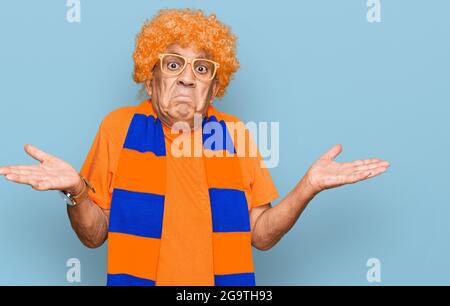 Senior hispanic man football hooligan cheering game clueless and confused expression with arms and hands raised. doubt concept. Stock Photo