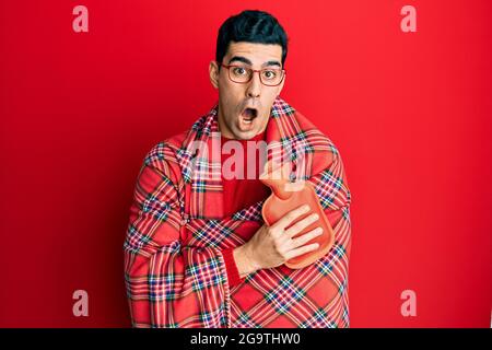 Handsome hispanic man wrapped in a winter blanket holding hot water bag afraid and shocked with surprise and amazed expression, fear and excited face. Stock Photo