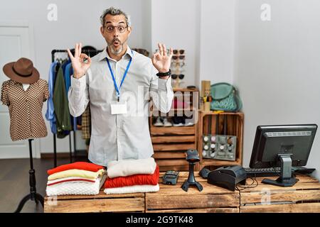 Middle age hispanic man working as manager at retail boutique looking surprised and shocked doing ok approval symbol with fingers. crazy expression Stock Photo