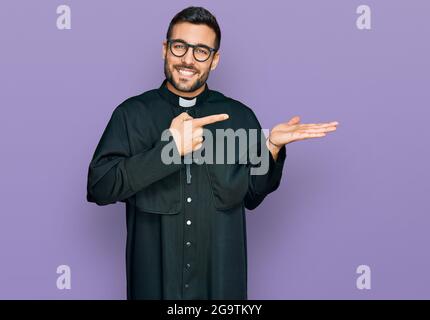 Young hispanic man wearing priest uniform amazed and smiling to the camera while presenting with hand and pointing with finger. Stock Photo