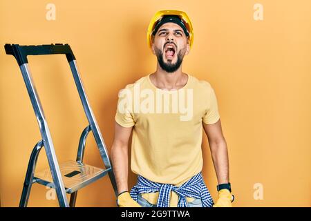 Handsome man with beard by construction stairs wearing hardhat angry and mad screaming frustrated and furious, shouting with anger. rage and aggressiv Stock Photo