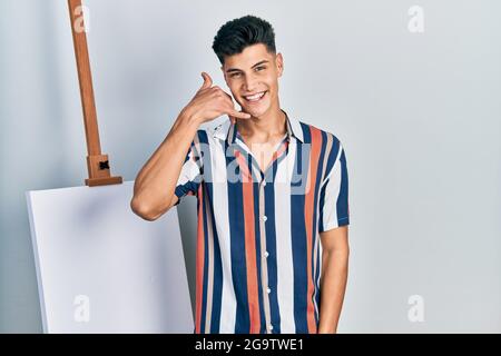 Young hispanic man standing close to empty canvas smiling doing phone gesture with hand and fingers like talking on the telephone. communicating conce Stock Photo