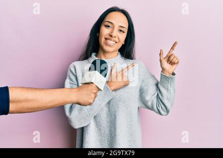 Young hispanic girl being interviewed by reporter holding microphone smiling and looking at the camera pointing with two hands and fingers to the side Stock Photo