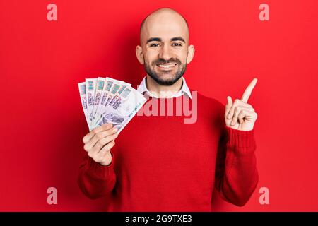 Young bald man holding egyptian pounds banknotes smiling happy pointing with hand and finger to the side Stock Photo