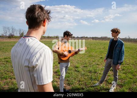 Young friends meet in a meadow in the open air with a guitar and sing songs - Male friendship and passion for music concept - focus on guitar Stock Photo