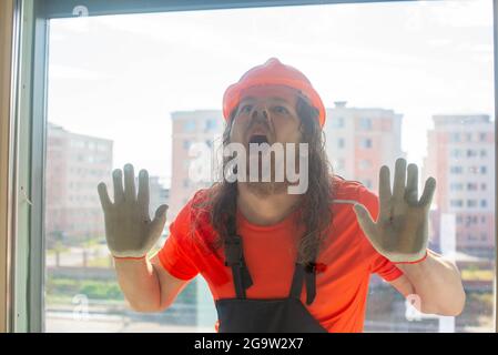 the long-haired engineer in the helmet makes a face behind the glass Stock Photo