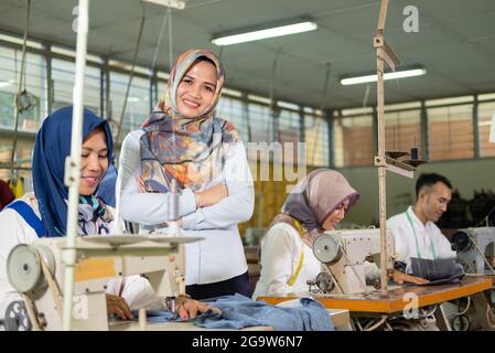 female supervisor in the headscarf smiling crossed hands stands to the side the seamstress using the sewing machine Stock Photo