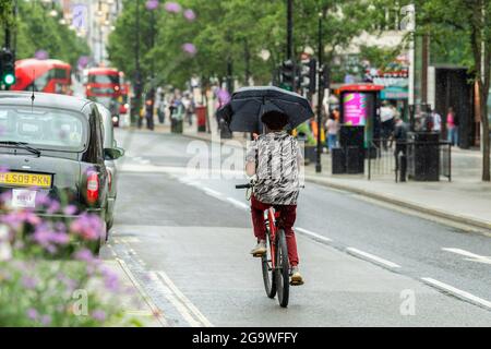 London, UK. 27th July, 2021. A man rides a bicycle along Oxford Street with an umbrella to protect him from the rain. Credit: SOPA Images Limited/Alamy Live News Stock Photo