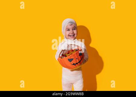 Child in mummy costume holding basket of chocolates in front of yellow background. Stock Photo