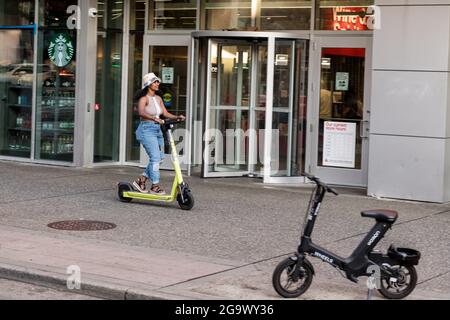 Seattle, USA. 27th Jul, 2021: A person on a ride share scooter on the sidewalk by target. The new scooter share scooters are not supposed to be ridden on sidewalks, but companies are also not required to provide helmets for riders to comply with local city helmet laws. This puts riders in a tough spot to either ride on sidewalks endangering pedestrians or ride in traffic with vehicles endangering themselves and therefore breaking the helmet laws. Credit: James Anderson/Alamy Live News Stock Photo