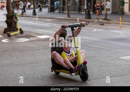 Seattle, USA. 27th Jul, 2021: A person on a ride share scooter on the sidewalk with a child by target. The new scooter share scooters are not supposed to be ridden on sidewalks, but companies are also not required to provide helmets for riders to comply with local city helmet laws. This puts riders in a tough spot to either ride on sidewalks endangering pedestrians or ride in traffic with vehicles endangering themselves and therefore breaking the helmet laws. Credit: James Anderson/Alamy Live News Stock Photo