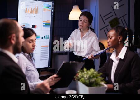 Diverse multi ethnic teamwork brainstorming company ideas analyzing financial graphs presentation. Businesspeople overworking in meeting room late at night discussing solution Stock Photo
