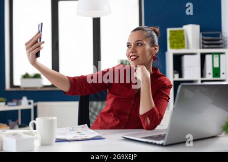 Successful businesswoman having fun at work taking selfies, sitting at desk office workplace using smartphone. Employee using phone camera to take photos of herself during break time. Stock Photo