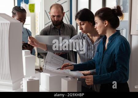 Team of diverse architects discussing blueprint plans on paper laptop computer tablet. Professional workers analyzing maquette building model for project design and construction Stock Photo