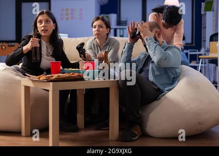 Team of multi ethnic friends losing tv game on console after work at office. Cheerful diverse group play with joystick while eating snacks pizza chips and drinking beer alcohol from cups Stock Photo