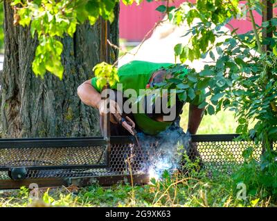 A male worker in a protective helmet welds metal using electric arc welding on a sunny summer day outdoors. Stock Photo
