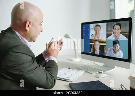 Mature businessman clapping hands while sitting at the table in front of computer monitor and talking to his colleagues during online meeting Stock Photo