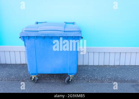 Single blue plastic trash can on wheels near building wall on asphalt Stock Photo